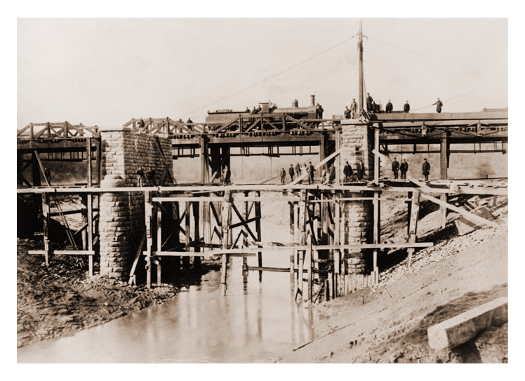The 'new' River Ely bridge with stone butresses is seen during construction with the original timber bridge to the rear.